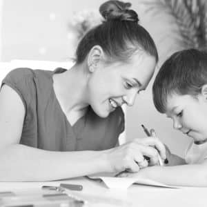 Horizontal shot of happy woman with hair bun, small boy, draw picture together, pose against domestic interior, have positive looks. Caring young mum spends free time with little son at home